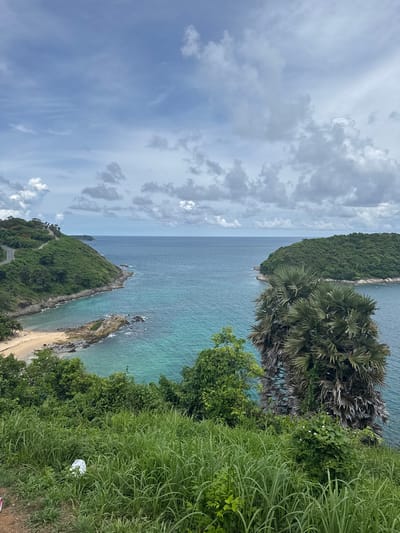 This is a photograph of a scenic coastal view in Rawai, Thailand. In the foreground, tall green grass covers a slope. In the middle ground, a bay with turquoise water is visible, featuring a sandy beach with a few people. Rocky outcrops are present along the coastline. To the left, a green, tree-covered hill rises, with a road winding its way up. To the right, another lush, green island juts into the sea, with a large palm tree in front of it. The sky is overcast with scattered clouds, and the sea is a deep blue.