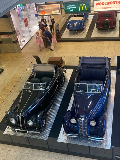 This media file shows an indoor scene, likely a shopping mall in Łódź, Poland, as indicated by the text "GALERIA ŁÓDZKA". Two vintage convertible cars are displayed on platforms. The top car is a dark blue convertible with its doors open, revealing a dark blue interior. The bottom car is a black convertible, also with its doors open. In the background, several people are gathered, including a group near a store window. One of the individuals is looking at their phone. A sign above them reads "Bezpłatny internet" (Free internet). Another sign visible in the upper left corner of the image reads "WOOLWORTH POZIOM -1" (Woolworth Level -1), suggesting a retail environment. There are also signs for "McDonald's" with "restauracja na poziomie +1" (restaurant on level +1). The lighting suggests it is daytime indoors. The overall atmosphere appears to be casual, with people browsing or observing the displayed cars and shopping.