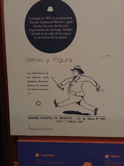 This image is a photograph of a museum exhibit about hats, specifically focusing on a Chilean hat shop called "Donde Golpea el Monito." The exhibit features a large, light-colored poster with dark blue text and a black and white illustration.
The poster at the top describes "Donde Golpea el Monito" as a hat shop founded in 1915, which is still one of the most important in Santiago, located on 21 de Mayo street. Below this text is a charming illustration of a man in a suit and hat, depicted in mid-stride, seemingly about to twirl a cane. The text on the poster also discusses the influence of a silhouette and how a good hat can enhance it. At the bottom of the poster, it is noted that this is a promotional leaflet from "Donde Golpea el Monito," dated 1937, from Santiago's National Library.
The exhibit also displays two actual hats. The first, labeled "1 CANOTIER," is described as being from around 1880 and made of braided straw from La Lajuela, Santa Cruz. The second hat, labeled "5 TRILBY," is from around 1890 and is made of merino wool felt.
The background of the photograph shows the dark blue wall of the exhibit space, with a section of a red border visible at the very bottom. The overall impression is one of historical display, celebrating fashion and the heritage of hat-making in Santiago.
