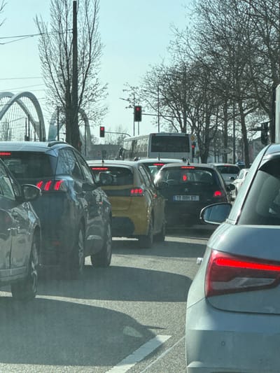 The image displays a multi-lane roadway in Kehl, Germany, under clear daylight conditions. A queue of vehicles is stationary, indicated by illuminated red brake lights. In the immediate foreground, the rear-right section of a light grey/silver car is visible, displaying an active red taillight. Directly ahead, a dark grey/black hatchback is stopped, bearing the French license plate "19 BBH 67" and active brake lights. To its left, a yellow hatchback with illuminated brake lights is positioned. Further left and slightly closer, a dark grey SUV exhibits distinctive illuminated red brake lights. A white bus is stopped further down the lane in the background. Multiple red traffic lights are visible on poles in the mid-distance. The background features leafless trees and the distinctive white arched structure of a bridge, consistent with the Europe Bridge. Shadows are cast onto the asphalt pavement from the vehicles and roadside elements. No pedestrians are visible.