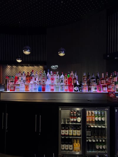 The image depicts a well-stocked bar display with numerous bottles of liquor and other beverages arranged on shelves. The bar appears to be located in the city of Manchester, United Kingdom, as indicated by the "TOILETS" sign in English. The bottles are illuminated from below by colored LED lights, creating a vibrant and attractive display. Behind the bar, a dark, textured wall is visible, and a few spherical pendant lights hang from the ceiling, casting a soft glow. A refrigerator unit is positioned below the bar, filled with various bottles and cans of drinks. The overall scene suggests a lively and modern bar or nightclub setting, likely busy with patrons, though no people are visible in the frame. The environment is clean and organized, with a focus on showcasing the beverage selection.