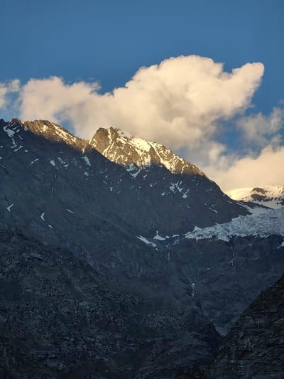 A majestic mountain range dominates the frame, its rugged, dark grey slopes covered in patches of snow. The peaks are jagged, catching the warm, golden light of what appears to be either sunrise or sunset, casting dramatic shadows across the landscape. A large, fluffy white cloud drifts in the bright blue sky, partially obscuring the sunlit summit. Below the prominent snowy peaks, a thick band of ice, likely a glacier, flows down the mountainside, its surface textured and uneven. The lower parts of the mountain in the foreground are covered in dark, rocky scree, adding to the raw, wild feel of the scene. The overall impression is one of a serene and awe-inspiring natural landscape, possibly taken in a mountainous region of India, given the location context. The lighting suggests a specific time of day, either early morning or late afternoon, with clear weather otherwise. There is no visible text in the image.