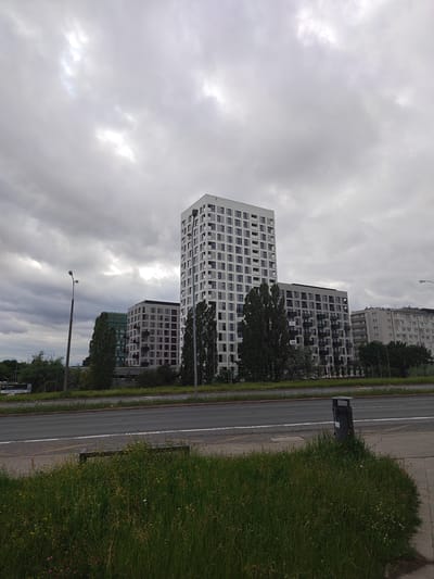 The image shows a modern residential complex in Poznan, Poland, on a cloudy day. The buildings are tall, with light-colored facades and numerous windows. Some buildings have balconies. In the foreground, there is a street with a sidewalk and a grassy area. A trash can is visible on the sidewalk. Trees are planted along the street and between the buildings. The sky is overcast with some patches of brighter light. A street sign is partially visible on the left side of the image. The scene appears to be a typical urban environment.