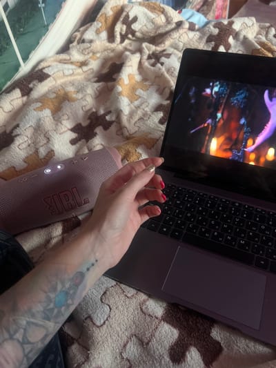 In Krasnoe-na-Volge, Russia, an individual's arm with tattoos is visible, resting on a pink JBL portable speaker. The speaker is positioned on a patterned blanket. A laptop with a movie playing on its screen is also present, its keyboard showing Cyrillic characters. The scene appears to be indoors, likely in a bedroom, with soft lighting and a comfortable, relaxed atmosphere.