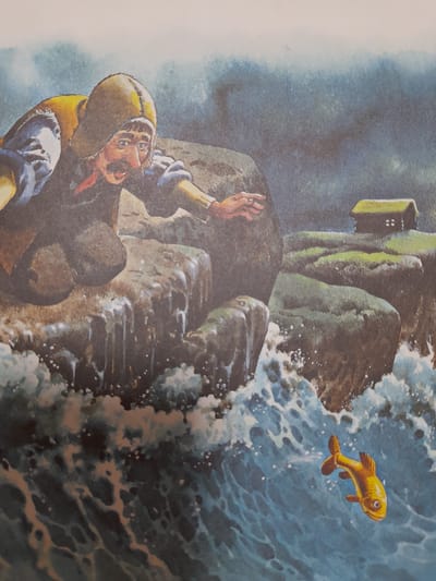 This image depicts a man, wearing a hooded jacket and with a mustache, kneeling on a rocky cliff overlooking the sea. His left arm is extended towards a small, golden fish swimming in the choppy blue water below. In the background, there is a small, dark structure with a green roof, possibly a house, situated on another rocky outcrop. The environment suggests a coastal setting with rough seas and overcast skies. The scene appears to be an illustration from a storybook or artwork. There is no indication of this scene being located in Baan Mae Nam, Thailand, or any specific city.