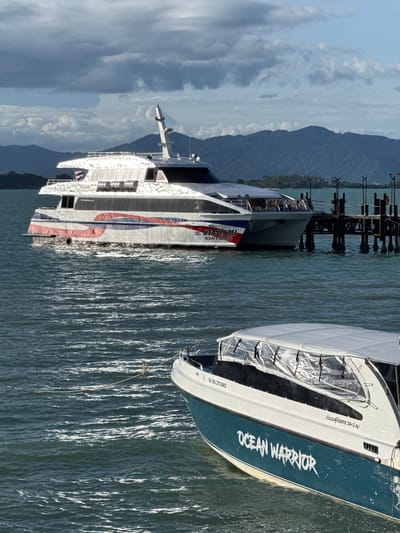 This is a wide shot of a ferry terminal at Baan Bang Rak, Thailand, with two boats in the foreground and a ferry docked behind them. The sky is overcast with patches of blue sky. The water is a deep blue with whitecaps. In the foreground, a white and teal speedboat named "OCEAN WARRIOR" is visible. The boat has a clear plastic canopy. Behind the speedboat, a large white and red ferry is docked at a wooden pier. The ferry has "KOH PRARB" written on its side in Thai and English. The pier has several lights and metal posts. In the background, there are lush green hills and trees.