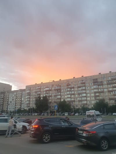 The image depicts a street scene in Telmana, Russia, featuring a large, multi-story apartment building on the right side of the frame. The building's facade is lined with numerous windows, suggesting a residential area. In the foreground, several cars are parked. One car, a dark sedan, is particularly prominent, its taillights illuminated, reflecting the warm hues of the sky. Another car, a white vehicle, is parked next to it, with a shopping cart nearby and a person standing beside it.
The sky dominates the left side of the image and displays a soft gradient of colors, transitioning from a pale pink and orange at the horizon to a muted gray and blue at the top. This suggests it is either sunrise or sunset. The presence of clouds indicates a cloudy or overcast sky.
A white van with Cyrillic text is visible parked behind some of the cars. The text on the van appears to be "ПЕТРОБАС" and a phone number. Traffic signs are also visible, including a blue directional sign with a white arrow.
The overall mood of the image is calm and somewhat serene, with the soft light of the sky creating a gentle atmosphere. The urban setting is clearly defined by the apartment building and the parked vehicles. The time of day is likely early morning or late evening due to the sky's coloration.