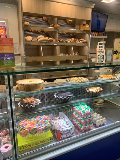 This image showcases a display case in what appears to be a bakery or confectionery shop in Odivelas, Portugal. The display is filled with a variety of baked goods, including loaves of bread, tarts, and individual cakes. Prominently featured are colorful lollipops arranged in a box labeled "LOOLY POP'S," as well as several packages of Kinder chocolate products. Various brands and product names are visible in the text, such as "Chupa Chups," "BEST&," "CLÁSSICO," "ORIGENS," "VITAL," and "COMPAL." The overall scene suggests a commercial establishment stocked with sweets and baked items. The lighting indicates it is daytime, and the orderly arrangement of products points to a well-maintained shop.