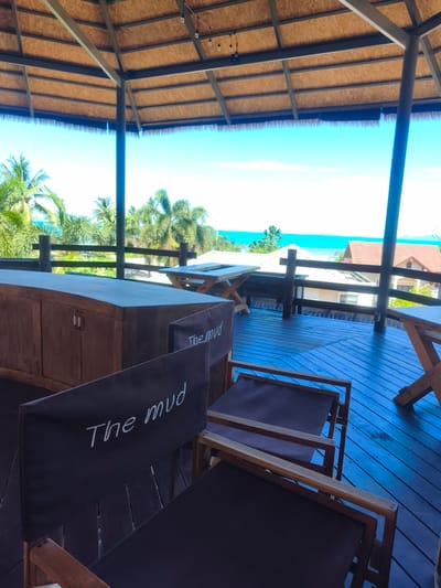 An outdoor covered seating area with a thatched roof overlooks the ocean in Baan Thong Po, Thailand. The foreground shows two dark canvas director's chairs with "The mud" written in white script on their backs. Behind the chairs, a curved wooden counter or bar is visible with two white drawer pulls. The floor of the seating area is made of dark wood planks. In the middle ground, there are several wooden tables with X-shaped legs and a wooden railing. Beyond the railing, palm trees and tropical vegetation are abundant, with houses visible in the distance. The bright blue ocean and sky form the background. The scene appears to be a scenic viewpoint or a restaurant/bar area. There are no people visible in the image.