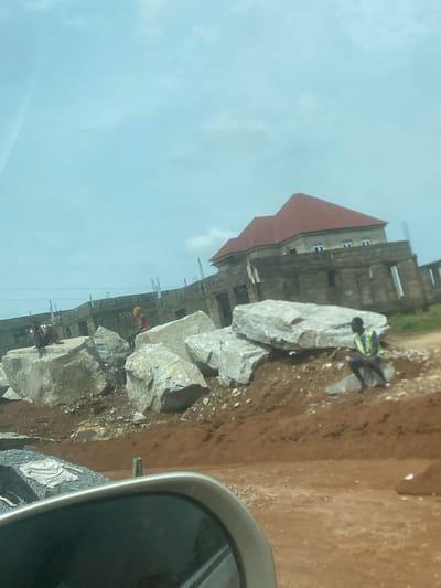 This image captures an active construction site in Gosa, Nigeria, under a bright, overcast sky. The perspective appears to be from inside a vehicle, with a portion of a car's side mirror visible in the lower right corner.
The central focus is a multi-story building in the early stages of construction, positioned on the left side of the frame. It features a prominent red-tiled roof at the top, while the main structure below consists of concrete and visible rebar. Several open window frames are visible on the building's facade.
In the foreground and mid-ground, a row of large, gray rocks of varying sizes runs across the scene, indicating construction materials. Several workers are present on the site. One worker, wearing a blue shirt and a high-visibility yellow-green vest, stands near these rocks. Further back, closer to the building, another worker is visible in a red or orange top, possibly wearing a hard hat. A third, less distinct figure can be seen near the base of the building on the left.
The ground is a mix of reddish-brown dirt and soil, characteristic of construction environments, with smaller debris scattered around. The bright, even lighting suggests it is daytime. No explicit text is clearly visible in the image.