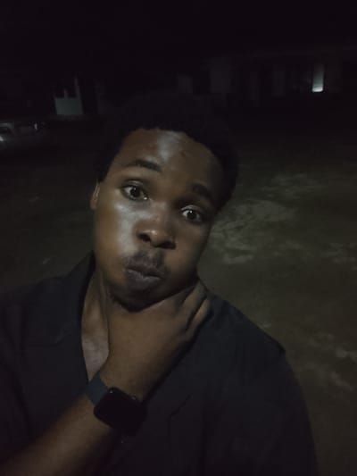 In Orodo, Nigeria, at nighttime, a dark-skinned young man with short black hair takes a selfie. He is wearing a dark collared shirt and a dark smartwatch on his left wrist. His right hand is touching his chin and neck. His mouth is pursed. The background is dark and appears to be an outdoor, possibly residential, area with faint structures visible in the distance. The lighting is low, casting shadows and creating a slightly eerie atmosphere.