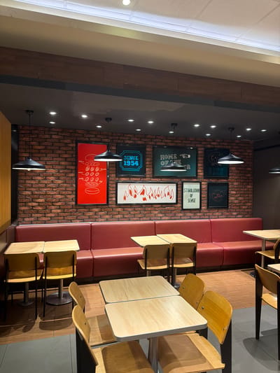 The image depicts the interior of a restaurant, likely located in the Jiangning District of China, evidenced by the inclusion of a phrase in Chinese that translates to "Originated in the United States, Popular around the World". The restaurant features a brick wall adorned with framed posters including one with the text "SINCE 1954" and "HOME OF THE ", another with a list of food components, and others with slogans. A built-in red bench seating runs along the wall adjacent to a row of wooden tables and matching chairs. Above the wall, a series of small spotlights are installed. The scene appears to be a quiet moment in the establishment, absent of people.