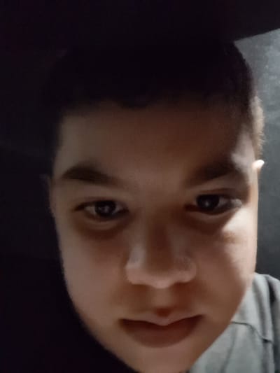 The image is a close-up, low-angle, poorly lit selfie of a young person, likely a male, with brown skin and dark hair. The individual is looking directly at the camera with a neutral expression. Their face is the primary subject, with their eyes, nose, and mouth visible. The background is dark and out of focus, suggesting an indoor setting. There is a hint of clothing with a striped pattern on the right side of the frame, possibly a shirt. The image does not contain any discernible elements that would allow for its location to be identified as Rustavi, Georgia.