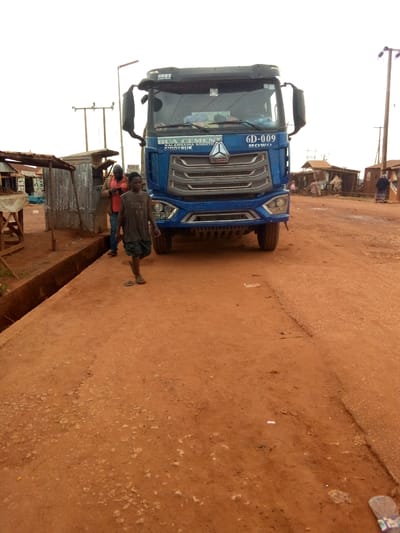 The image depicts a daytime scene on an unpaved, reddish-brown dirt road, likely in a rural or semi-urban area of Mbar, Nigeria. A large blue Sinotruk HOWO heavy-duty truck is prominently featured, facing the viewer and occupying much of the central and right foreground.
On the truck's windshield, a banner reads "BUA CEMENT KALAMBAINA SOKOTO SINOTRUK," while "6D-009 HOWO" and "中国重汽 SINOTRUK" are visible above its grill. A young boy walks towards the viewer on the dirt road in front of the truck, wearing a faded gray t-shirt with partially visible text, possibly "UNFLU," and dark shorts, with one sandal on and the other off.
To the left, behind the boy, another man stands near simple structures made of corrugated iron, which appear to be market stalls or small shops. This man is wearing an orange shirt and blue jeans. A dark, open drainage ditch runs along the left side of the road. A blurry sign partially visible behind the man seems to say "FREEDOM ART."
The sky is bright, suggesting either an overcast day or strong midday sunlight. In the background, power poles with multiple lines are visible against the bright sky, and more distant buildings and figures can be discerned. The road surface is uneven, consistent with an unpaved track, and shows some scattered debris. The overall scene captures a moment of daily life and commerce in the area.