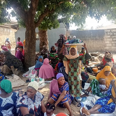 An outdoor scene in Doma, Nigeria, displays a significant gathering of women and children on dusty ground beneath a large tree with green foliage. The majority of individuals are seated, many on fabrics spread on the ground, dressed in varied traditional garments and head coverings. A young female stands prominently in the center, balancing a silver-colored metal tray laden with folded textiles on her head, her arms slightly raised. Various bags, bundles, and an orange bucket are scattered on the ground around the seated individuals. In the background, structures built from concrete blocks are visible, with a building featuring a corrugated iron roof further in the distance, under bright daylight conditions.
