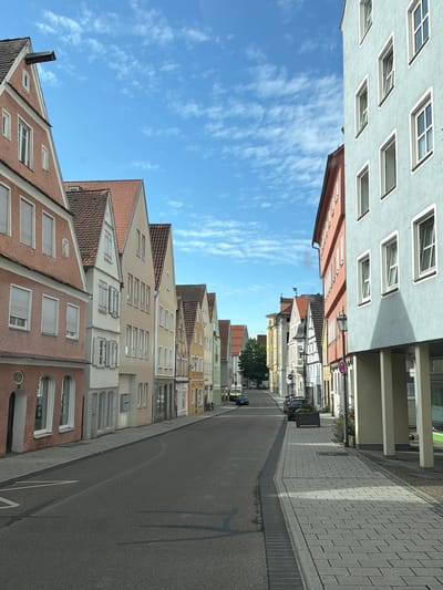 The image shows a street scene in Memmingen, Germany, with a row of colorful buildings on the left and a modern building with light blue facade on the right. The street is paved with asphalt and lined with a sidewalk made of grey paving stones. On the left side of the street, there are several old buildings with pitched roofs and various colored facades, predominantly in shades of pink, yellow, and cream. These buildings have traditional windows and some have shutters. On the right side of the street, a contemporary building with a light blue facade stands out. This building has multiple windows and is supported by large white pillars, creating a covered walkway. The sky is blue with scattered white clouds. Several cars are parked along the street and on the sidewalk. In the background, more buildings and trees are visible, suggesting a European town or city setting. The scene appears to be captured during daylight hours, under a clear sky.