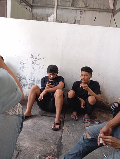 The image depicts a casual gathering of at least four men in what appears to be a utilitarian or informal concrete space in Katapang, Indonesia. Two men are prominently seated on the concrete floor, leaning against a weathered white wall with visible cracks and peeling paint.
The man on the left is wearing a black t-shirt, black shorts, a black baseball cap, and black flip-flops. He has a white patch or mask under his nose and is engrossed in his smartphone, which is connected to white wired earbuds. He holds a cigarette in his left hand.
Next to him, the man on the right is wearing a black t-shirt, ripped black pants, and blue flip-flops. He has short dark hair, ear gauges, and a light mustache. He is holding a lit cigarette to his mouth with his right hand and another unlit cigarette in his left. A small clear plastic cup containing a dark liquid and a cigarette pack are visible on the ground near him.
Partial views of other individuals are seen at the edges of the frame. On the far left, a person's shoulder, arm (in a light blue shirt), and legs (in light blue ripped jeans and sneakers) are visible. On the far right, legs in ripped blue jeans and an arm holding a lit cigarette are seen. The group appears to be socializing or taking a break. Most of the visible individuals are smoking cigarettes.
The setting includes a dirty concrete floor and an unfinished ceiling structure with beams and more cracked concrete above the wall. The lighting suggests it is daytime, casting a soft, ambient glow on the scene. The overall atmosphere is relaxed and informal. No discernible text is visible on clothing or surrounding objects.