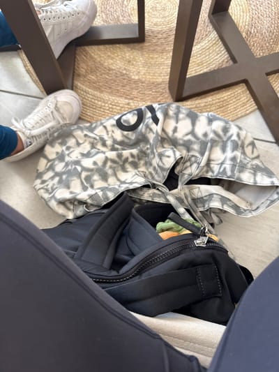 This low-angle, downward shot captures a person's lower body and belongings on the floor, likely taken from a seated position. The primary subjects are the person's legs and feet, clad in white sneakers. One leg, wearing dark leggings or athletic pants, is prominent on the right, while the other, wearing blue pants, is partially visible on the left.
Two bags rest on the light-tiled floor. On the left is a large, off-white tote bag featuring a grey, abstract, tie-dye or camouflage-like pattern. A large black "O" or "0" is distinctly visible on its side. To the right of the tote bag is a black backpack or duffel bag with a prominent silver zipper.
The setting appears to be an indoor environment, possibly a cafe or restaurant in Puebla City, Mexico. Dark brown, angular table legs are visible in the foreground, forming a geometric pattern. A circular, woven natural-fiber rug is partially visible beneath the table legs and feet, adding texture to the floor. The scene depicts a casual moment, with bags placed conveniently on the floor while the person is seated. The time of day, weather, and emotions are not discernible from the image.