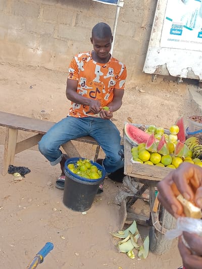 A man wearing a TikTok t-shirt sits on a wooden bench outside, peeling fruit with a small knife. A black bucket overflowing with peeled fruit sits between his feet. To his right, a wooden cart displays watermelon slices, oranges, and bananas. Peels of fruit and other debris scatter the ground. A sign is visible behind him, advertising President Buhari. A person's hands are peeling fruit in the foreground.