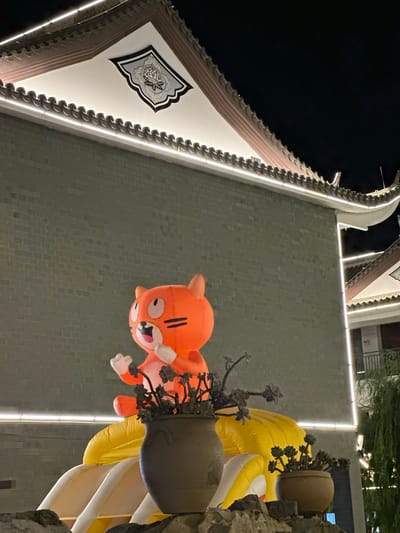 An outdoor nighttime scene depicts a large, inflated cartoon tiger character with an orange body, white paws, and black facial features, appearing to be on a yellow inflatable structure. The tiger is positioned in front of a grey brick building with traditional East Asian architectural elements, including a curved, tiled roof illuminated by white LED lights. Several potted plants are placed in the foreground, obscuring the lower portion of the inflatable structure and the tiger's body. The background features more of the illuminated building and the dark night sky. The location is identified as Eshan Yi Autonomous County, China. There are no people visible in the image.