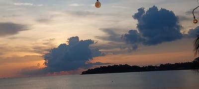 A wide-angle shot shows a tranquil scene at Baan Sri Thanu, Thailand, at dusk. The sky features a blend of orange, yellow, and blue hues, with scattered clouds. Two dark, dense cloud formations dominate the skyline. A landmass silhouetted with trees stretches across the middle ground, bordering the calm water in the foreground. Above, two incandescent light bulbs are suspended from wires. Palm fronds partially intrude into the frame on the right. The water is still with very few objects in the water.