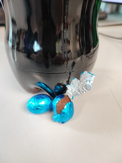 The image displays a close-up, ground-level view of the base of a dark, reflective plastic bottle, likely a beverage container, situated on a light-colored, flat surface. The bottle's surface exhibits distorted reflections of the surrounding indoor environment, suggesting light sources and possibly other objects. Positioned next to the bottle's base are three small, egg-shaped chocolate candies. One candy is fully encased in bright blue metallic foil. Another, located centrally, is partially unwrapped, revealing milk chocolate beneath torn and crumpled blue foil, with silver foil visible around the exposed chocolate. A third blue-foiled candy is partially visible behind the unwrapped one. The scene is located in Obruchishte, Bulgaria.