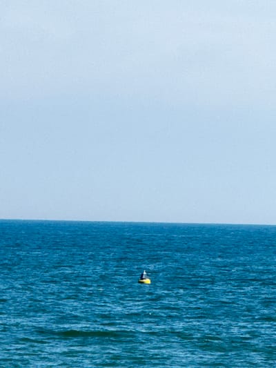A single yellow buoy with a black top floats on the deep blue ocean under a pale blue sky. The horizon line is clearly visible, separating the water from the sky. There are no people or other objects in the image.