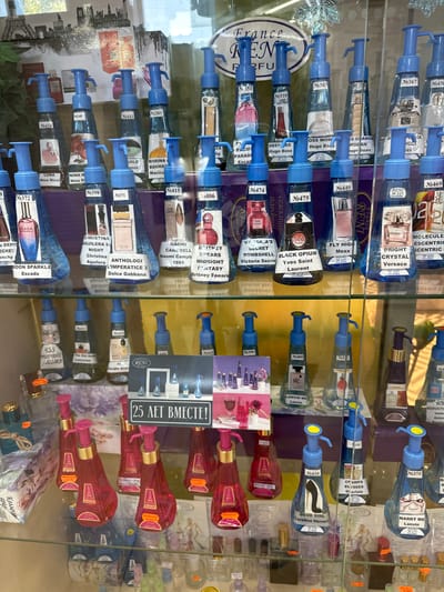 This image depicts a retail display of perfume bottles, likely in a shop in Novoaltaysk, Russia. The perfumes are arranged on shelves behind a glass or plastic barrier. The bottles are mostly clear and filled with colored liquids, topped with blue pump dispensers. Each bottle has a label with a number, brand name, and sometimes a scent description.

In the foreground, several rows of these perfume bottles are visible. The top two rows feature bottles with blue dispensers, while the bottom rows showcase bottles with pink pump dispensers, some with gold accents. There are also some smaller bottles and decorative elements.

A sign with Cyrillic text and the words "France REM PARFUM" is prominent in the upper portion of the display. Another sign in the middle of the image reads "25 ЛЕТ ВМЕСТЕ!".

The background shows what appears to be a wall display with images of iconic Parisian landmarks, such as the Eiffel Tower. The lighting suggests an indoor retail environment, possibly a perfume or cosmetics store. No people are directly visible in the image, but the presence of a retail display indicates a commercial setting.