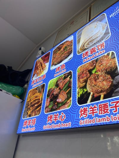 This media file displays a brightly lit, backlit menu board, likely from a restaurant or street food stall in Guang Zhou Shi, China. The board features a vibrant blue background with a circular pattern and showcases six different food items with accompanying photographs, Chinese names, and English translations.

The featured dishes include:
1.  **烤囊饼 (Baked Cake):** Illustrated by a stack of round flatbreads.
2.  **烤羊腰子 (Grilled Lamb Loin):** Pictured as chunks of grilled lamb on skewers, seasoned with herbs. The English translation is partially visible as "Grilled lamb loi".
3.  **烤鱼 (Roast fish):** Shows a whole roasted fish, garnished with green herbs.
4.  **烤羊心 (Grilled lamb heart):** Depicts skewers of grilled lamb heart, seasoned and charred.
5.  **烤羊肉串 (Shish kebab):** Features multiple skewers of grilled meat, a common street food item.
6.  **烤羊排 (Rack of Lamb):** Displays a rack of grilled lamb ribs, cooked to a golden hue.

The menu is designed for clear presentation, making it easy for customers to identify and choose dishes. A small white device, possibly a security camera or sensor, is visible below the menu board on the left side. The backlit nature of the display suggests it might be used in an indoor setting or during the evening/night.