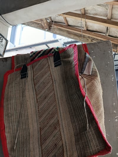 An outdoor scene in Depok, Indonesia, shows two woven mats hanging from a concrete ledge under a wooden roof structure with exposed wiring. The mats, one predominantly tan with red trim and diagonal brown stripes, and another visible at the top with striped green, pink, and black trim, are hung by black fabric loops and thin gray cords. A white and grey fabric tag is attached to the right mat. The background reveals a bright sky and a glimpse of a building through an opening in the roof. A partially visible sign with the number "8" and text in a foreign language is on the far left.