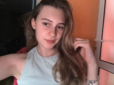 A young woman with long, wavy brown hair is shown in a close-up, frontal, selfie-style shot. She is wearing a light blue ribbed tank top and a silver necklace with a small heart-shaped pendant. Her left arm is bent, with her hand resting near her head, and her right arm is visible on the right side of the frame, adorned with a silver charm bracelet. She has fair skin, brown eyes, and is looking slightly off to her left. The background consists of an orange textured wall on the right and a dark surface on the left. A sliver of a white door frame with a handle is visible on the far right. The image does not provide any specific context that would allow for its location in Panagyurishte, Bulgaria.