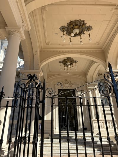 The image shows the interior of a building in Tigre, Argentina, notable for its ornate architecture. The perspective is looking upwards from what appears to be a balcony or landing with wrought-iron railings. The building features cream-colored walls with elaborate moldings, decorative wall sconces with multiple light bulbs, and an arched design overhead. A large column supports the structure. Windows are visible beyond the balcony railings. The building exhibits a classical architectural style.