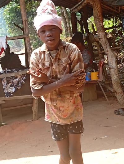 A young individual stands outdoors, wearing a pink head covering, a tan plaid shirt over a patterned undershirt, and black shorts. Arms are crossed, hands gesturing. Behind, a weathered wooden structure frames the scene with miscellaneous items like chairs, buckets, and clothes visible. The ground is reddish-brown. Another person is partially visible in the background, seated near the wooden structure.