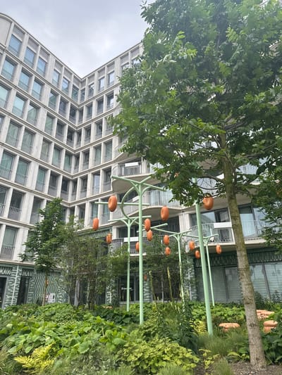 The image depicts a modern building with a curved facade constructed with light-colored bricks and numerous windows with balconies. In the foreground, there is a lush green garden with dense foliage. Several decorative elements resembling large orange pods are suspended from light green, tree-like structures. A mature tree with a textured trunk stands to the right of the frame, its branches laden with green leaves. The scene is set against an overcast sky. The building appears to be an apartment complex or office building, likely in a contemporary urban development. Research indicates this scene is likely located in Amsterdam, Netherlands, specifically at the Amsteldok building, a mixed-use development known for its distinctive architecture and integrated green spaces.