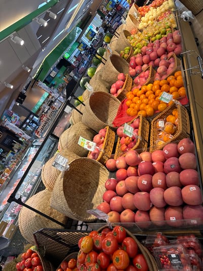 Here is a description of the image:
The image is a high-angle shot taken in a supermarket, likely located in Kerobokan Kelod, Indonesia. The main focus is a fruit and vegetable display section. Baskets made of woven material hold various produce, including apples, oranges, pears, and tomatoes. The fruits and vegetables are neatly arranged, creating a visually appealing display. Price tags are visible, indicating the cost of some of the items.
In the background, other supermarket aisles are visible with a variety of products on shelves. The lighting is bright, typical of a retail environment. Signage in both English and Indonesian are seen. One sign promotes "Japanese Cuts" for Yakiniku, Sukiyaki, and Shabu Shabu.