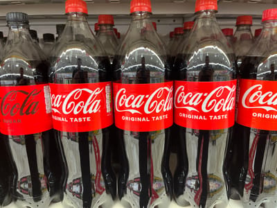 The image shows a close-up of several plastic bottles of Coca-Cola Original Taste on a shelf, likely in a supermarket. The bottles are mostly filled with the dark brown liquid, and their red labels with the Coca-Cola logo are clearly visible. Some bottles have red caps, while one on the far left has a black cap.
The labels contain text, including "Coca-Cola Original Taste," and nutritional information such as "0 SUGAR" and "0 CALORIES" on one bottle. There is also a best-before date visible on several labels, such as "26/08/2026" and "27/08/2026," along with lot codes.
Reflections on the plastic bottles show a blurry image of a person taking the photograph, with overhead lighting fixtures reflected in the beverage. The overall impression is a commercial display of a popular soft drink. The location context is given as Kharkiv, Ukraine, which suggests the setting is a retail store within that city. There are no indications of weather, time of day, or specific emotions conveyed, other than the neutral presentation of merchandise. The visible text is primarily product labeling and manufacturing information.