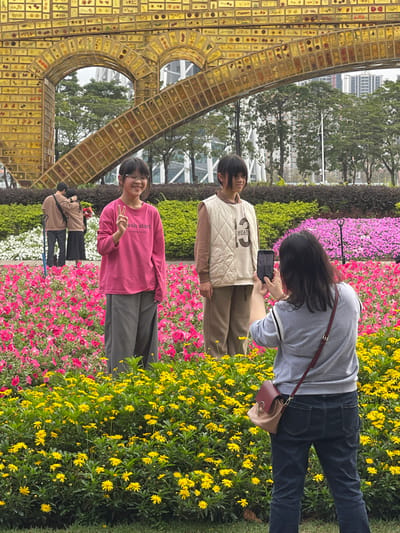 The image captures three young women posing for a photo in a garden in Guangzhou Shi, China, located at the coordinates 23.115 latitude and 113.321 longitude. The women are standing in a bed of vibrant pink and yellow flowers, with the tallest woman in the back wearing a gray sweater and dark jeans. She is holding a brown purse with a gold chain, and she appears to be taking a selfie with her phone. The middle woman is wearing a beige quilted vest over a white shirt, and she has her hands clasped in front of her. The shortest woman is standing at the front, wearing a pink shirt and a pair of khaki pants. She has one hand raised, as if in a playful gesture. The three women are smiling at the camera. Behind them is a large, gold-colored structure, possibly an art installation or a building, with a textured surface. The flowers in the background are arranged in a seemingly organized manner, creating a visual contrast between the vibrant colors and the structure's smooth surface. The scene is bathed in natural light, and the overall mood is cheerful and bright.