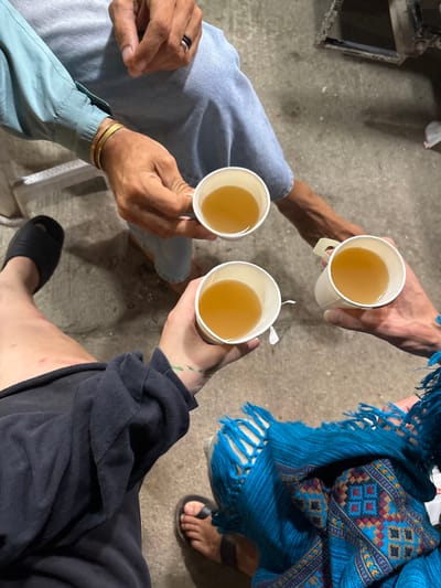 Three individuals raise paper cups filled with a golden-colored liquid, possibly tea. The person on the left wears a blue button-down shirt and denim pants, adorned with a gold bracelet and a black ring. The person in the middle is dressed in dark clothing and wears black sandals. The person on the right is wearing a blue patterned shawl, also wearing black sandals. Their feet are visible on a concrete floor.
