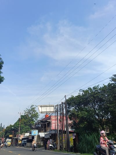 A daytime street scene features a row of small shops or eateries lining the road. People are gathered outside one of the establishments. Power lines crisscross the clear blue sky with sparse clouds. Vehicles, including motorcycles and a small truck, move along the road. Lush green trees frame the buildings. A sign extends from one of the structures.
