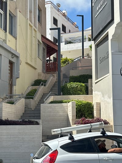 The image shows a street scene in Paphos Municipality, Cyprus, featuring a multi-level outdoor staircase adjacent to buildings. The staircase is constructed of concrete with metal railings and is flanked by planters with lush green foliage and some reddish-purple plants. On the right side of the image, a white car is visible in the foreground, partially obscuring the lower part of the staircase. A person wearing a hat is seated in the driver's seat of the car, looking to the right. The car has a white cylindrical object strapped to its roof rack. In the background, several buildings of varying architectural styles are visible, including white structures with balconies and a building with a light yellow facade. A dark lamppost stands near the staircase. A sign with text that reads "Sotris" is mounted on the building to the right. The sky is clear and blue. The overall scene depicts a sunny day in a developed urban environment with a focus on the architectural elements and vegetation.