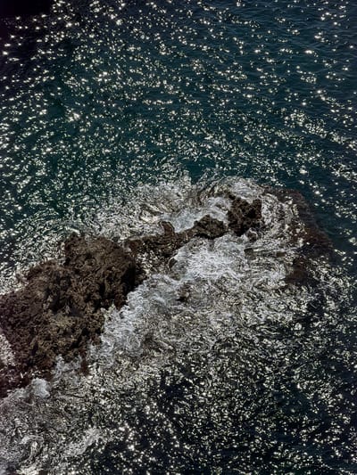 An overhead shot depicts rocky formations where dark blue water meets and creates whitewater. The water reflects light in a sparkling pattern, and the rocky area interrupts the water's surface. The location is near Prijevorac, Montenegro.