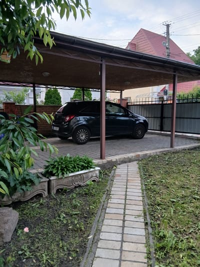 This image depicts an outdoor scene at a residential property in Vita-Poshtova, Ukraine. A black hatchback car is parked under a dark brown carport with a corrugated roof. The carport is supported by brown metal posts and sits on a paved area constructed with interlocking bricks. Adjacent to the carport is a grey metal fence. In the foreground, a path of light-colored pavers runs through a grassy area with some small plants and two decorative concrete planters. Lush green foliage from a tree overhangs the left side of the frame. In the background, a house with a red tiled roof and a utility pole with electrical wires are visible. The sky is overcast. No people are present in the image.