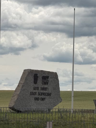The image shows a granite stone monument with black lettering and a crest. The text on the monument reads "OMV GUTE FAHRT! STADT SCHWECHAT UND OMV". This monument is located in the city of Schwechat, Austria, and it commemorates the city and the OMV company. The monument is situated in an open field with a cloudy sky in the background and a metal fence in the foreground. A tall white flagpole stands behind the monument. The overall scene suggests an outdoor location, possibly a roadside or an entrance to a facility.