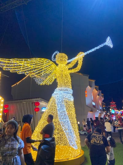 The image depicts a vibrant nighttime scene at Kya-Kya Kembang Jepun, Surabaya, Indonesia, illuminated by extensive festive decorations. A large, prominent wireframe sculpture of an angel blowing a trumpet stands centrally, constructed from myriad golden-yellow and white lights. The angel features golden wings and body, with white lights forming the trumpet, a diagonal sash across its body, and a halo.
In the foreground, a young female with light-colored hair and a light blue headband is visible on the left, wearing a patterned dress. Adjacent to the angel's base on the left, a person wearing an orange shirt and a black hijab is seen from behind, standing near a male in a grey shirt with a red armband, who has his arms crossed and is holding a black trash bag.
The background reveals architectural structures typical of East Asian design. On the right, a building with a multi-tiered roof and visible signage reading "KYA-KYA" is present. Further right, a red pagoda-like structure can be seen. On the left, another building features vertical white paneling and several suspended red spherical lanterns. Numerous other individuals, comprising adults and children, are distributed throughout the mid-ground and background, observing the decorations and moving across what appears to be a paved area combined with sections of artificial green turf. The sky is dark blue, indicating the night environment, which is brightly lit by the decorative installations.