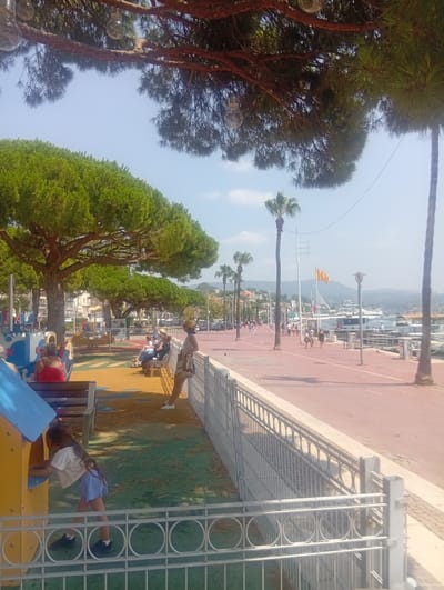 The image depicts a sunny day scene along the waterfront in Bandol, France. In the foreground, a children's playground area is visible with a yellow and blue structure and a girl playing. To the right, a metal fence separates the playground from a red-paved promenade. Several people are seated on benches in the middle ground, observing the surroundings or resting. The promenade is lined with palm trees and leads to a harbor filled with boats and yachts. Buildings can be seen in the background, suggesting a town or city setting. The sky is clear blue with a few wispy clouds. The overall atmosphere is one of leisure and a pleasant summer day.