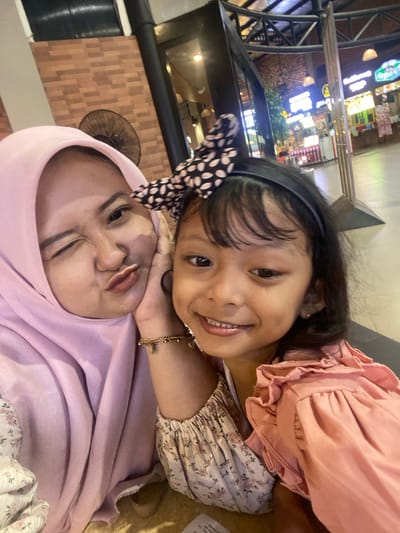 A selfie-style portrait captures a woman in a pink hijab and a young girl with a polka-dot bow in her hair. They are positioned close together. The background indicates an indoor setting, possibly a restaurant or mall in Surabaya, Indonesia, as suggested by the building features and lighting.