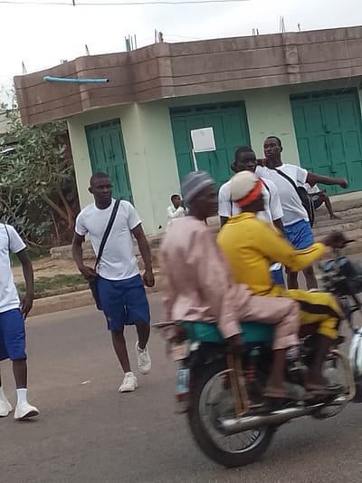 In Pantame, Nigeria, a group of young men in white t-shirts and blue shorts walk along a paved road. One man in the foreground carries a black bag across his chest, while another man behind him points to the right. Further in the background, a motorcycle carries two passengers: one in a light brown outfit and another in a yellow jacket, both wearing head coverings. The motorcycle is in motion, creating a blur effect. Behind the pedestrians, a light green building with ornate brown trim and closed doors stands under a cloudy sky. The scene appears to be a casual street encounter on an overcast day.