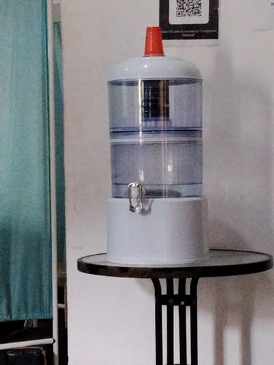 A water dispenser with a blue filter housing and a clear water reservoir is placed on a small round table. The dispenser has a bright red cap on top. The table has a dark, possibly glass or metal, surface and a black, multi-legged stand. To the left of the dispenser, a teal-colored curtain is visible. In the background, a white wall is present, with a black and white QR code affixed to it. The scene is indoors, likely in a domestic or office setting. The lighting appears to be artificial and ambient. No people are visible in the image. The time of day is not discernible. The location is Gwarinpa, Nigeria.