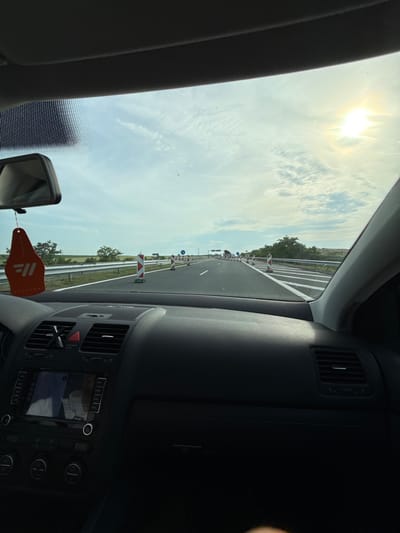 The image shows a view from inside a car driving on a highway. The road is straight and has orange and white construction barriers and signs along the side, indicating possible road work. The sky is bright with the sun visible. The dashboard of the car is visible, with air vents, a navigation screen, and controls. An air freshener hangs from the rearview mirror. The location is likely near Hadzhidimitrovo, Bulgaria based on the navigation on the screen.