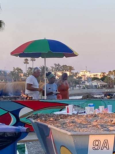 In Marbella, Spain, on a beach, a group of people are gathered around a colorful beach umbrella. An older man in a white t-shirt stands to the left, holding a plate and looking at a barbecue. Beside him, a person wearing a blue shirt and a head covering holds a plate. To the right of the umbrella, a woman in an orange dress is visible. In the foreground, a brightly painted boat or structure is present, with several white cups, a Coca-Cola bottle, and other items on a surface behind it. The background shows palm trees and buildings, suggesting a coastal resort area. The scene appears to be set during an evening gathering, possibly a barbecue or a meal on the beach.